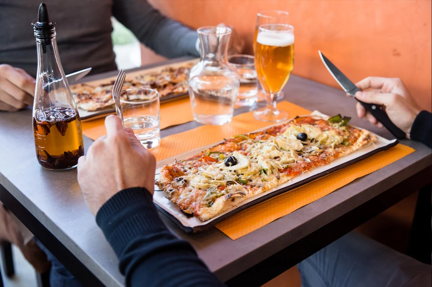 Deux convives trinquent autour d'une Long Pizza®, bière blonde et carafe posées sur la table