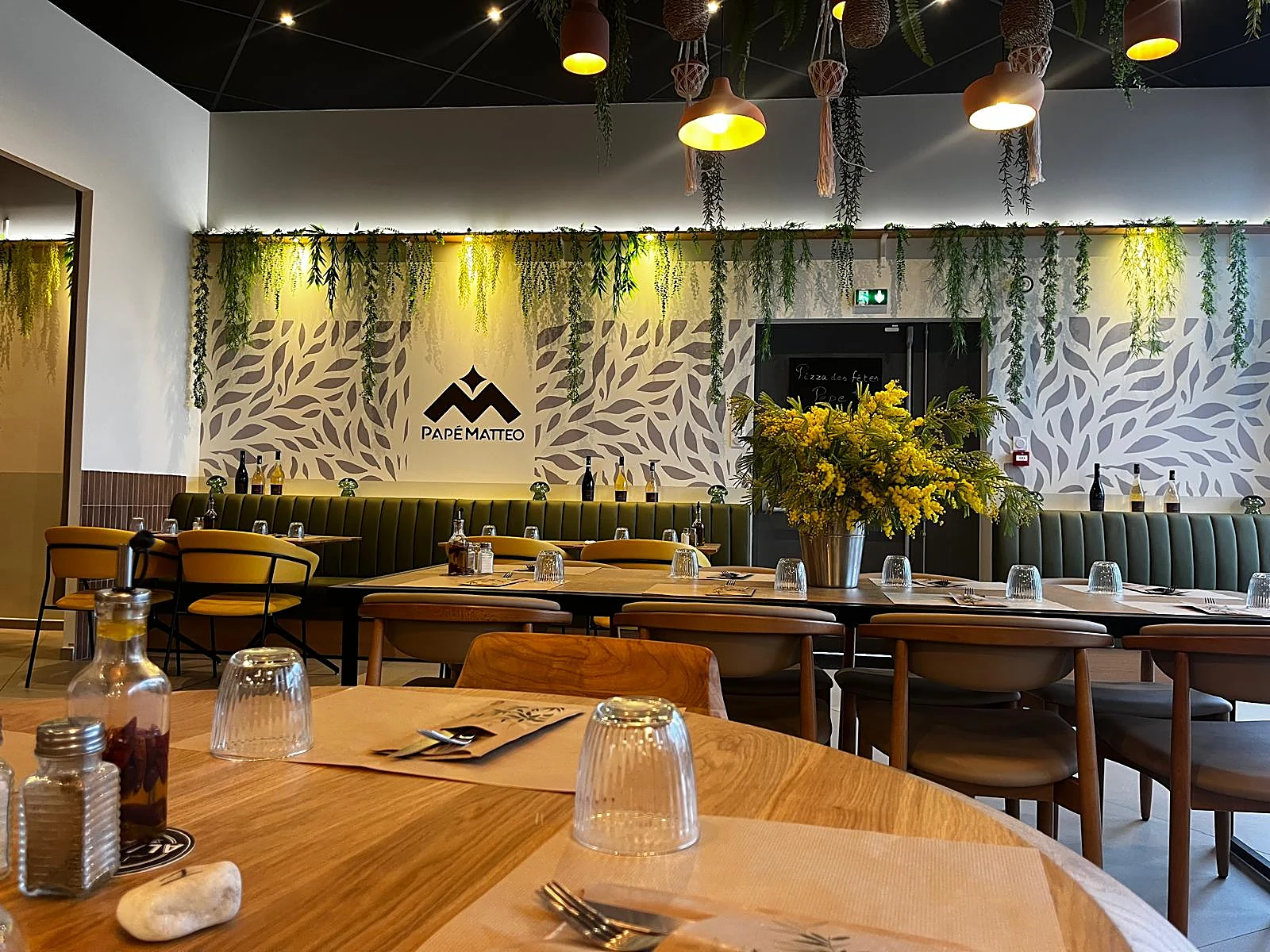 La salle de Lodève dressée pour le service : table ronde au premier plan, banquettes vertes, mur au feuillage et grand M du logo au fond